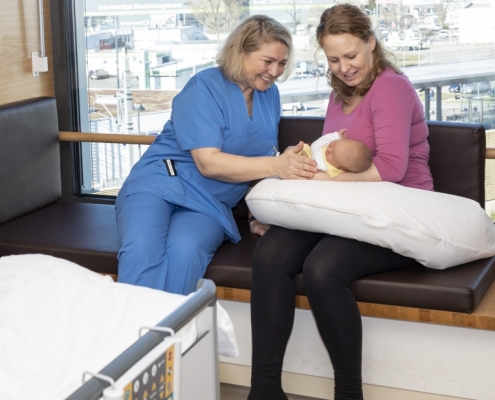 Mutter sitzt mit Neugeborenem und Pflegerin vor einem Fenster